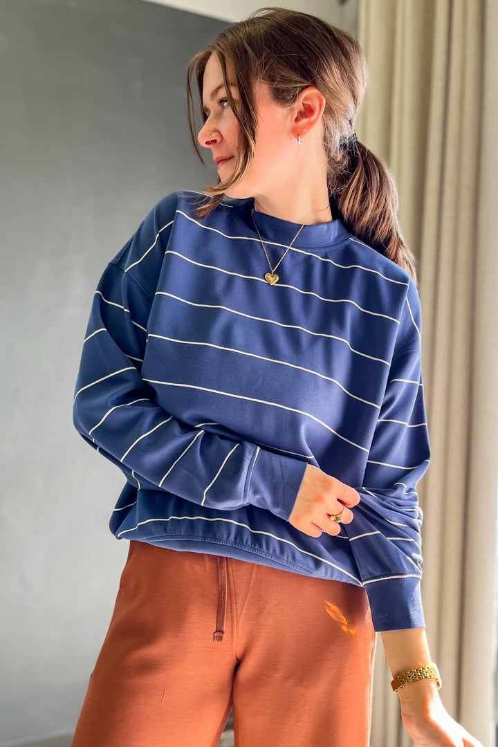 A woman wearing a blue long-sleeve sweatshirt with thin white horizontal stripes and rust-brown drawstring sweatpants, standing indoors against a gray wall and beige curtain.