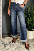 A person wearing dark blue wide-leg cuffed jeans with a faded wash, paired with brown woven slip-on shoes, standing indoors on a patterned rug next to a potted plant.