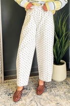 A person wearing white wide-leg cropped pants with a black polka dot pattern, styled with a yellow striped sweater and brown woven sandals, standing indoors next to a potted plant.