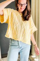 A woman wearing a pale yellow cable-knit short-sleeve top and light blue jeans, standing indoors with a neutral background.