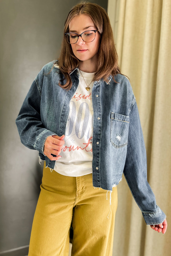 A woman wearing a light-wash blue denim jacket over a white graphic t-shirt and mustard yellow pants, standing indoors against a neutral gray and beige background.