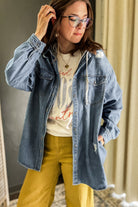 A woman wearing a light blue oversized denim shirt jacket over a cream graphic t-shirt and mustard yellow pants, standing indoors against a neutral background.