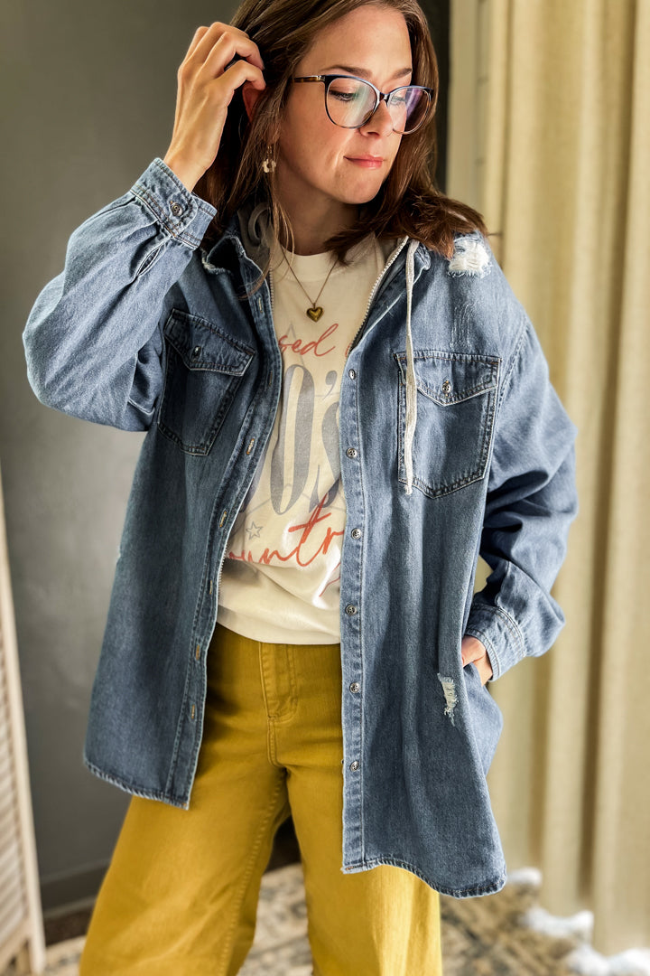 A woman wearing a light blue oversized denim shirt jacket over a cream graphic t-shirt and mustard yellow pants, standing indoors against a neutral background.