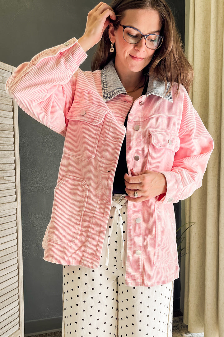 A woman wearing a pink corduroy jacket with a denim collar, styled with white polka dot pants and a black top, standing indoors.