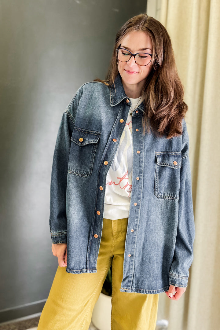 A woman wearing a faded blue denim button-up shirt jacket over a white graphic tee and mustard yellow pants, standing indoors against a dark gray wall and beige curtain.