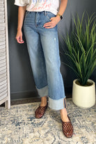 A person wearing light blue cropped jeans with rolled cuffs, paired with brown woven flat shoes, standing indoors against a dark gray wall next to a potted green plant.