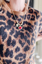 A close-up of a person wearing a gold chain necklace with three small oval charms, including a prominent red oval charm, over a brown and black leopard-print knit sweater.