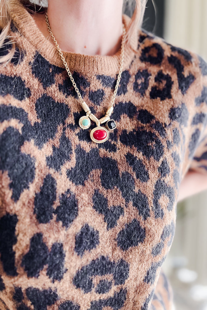 A close-up of a person wearing a gold chain necklace with three small oval charms, including a prominent red oval charm, over a brown and black leopard-print knit sweater.