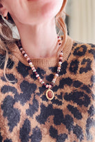 A close-up of a woman wearing a beaded necklace with a gold-toned oval pendant over a brown and black leopard print sweater.