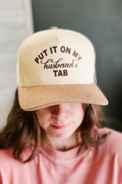 A person wearing a beige and brown ribbed trucker cap with the text 'PUT IT ON MY husband's TAB' in brown lettering, shown in a close-up lifestyle shot.