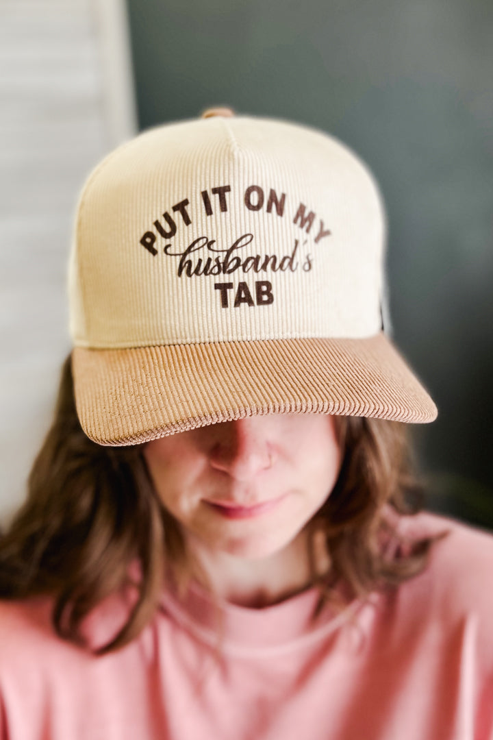 A person wearing a beige and brown ribbed trucker cap with the text 'PUT IT ON MY husband's TAB' in brown lettering, shown in a close-up lifestyle shot.