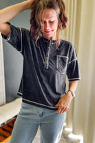 A woman wearing a black washed short-sleeve henley shirt with white contrast stitching and light blue jeans, standing indoors near a window with curtains.