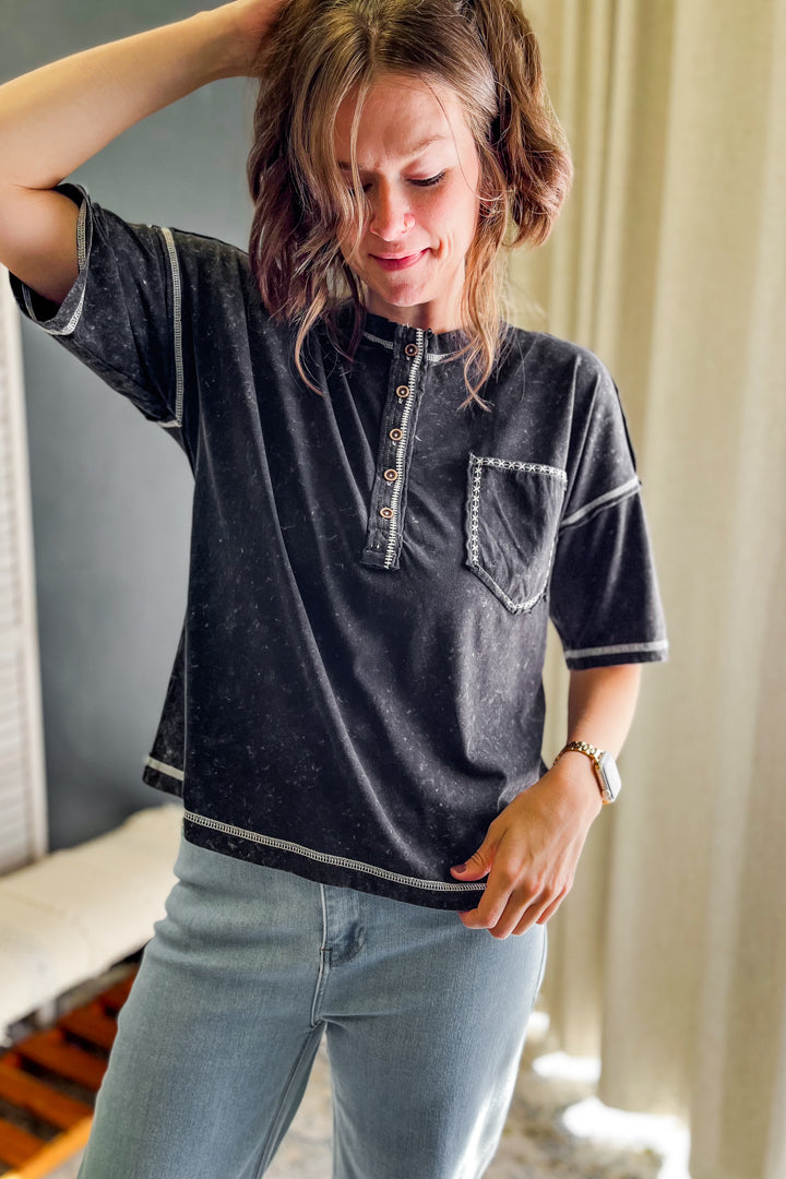A woman wearing a black washed short-sleeve henley shirt with white contrast stitching and light blue jeans, standing indoors near a window with curtains.