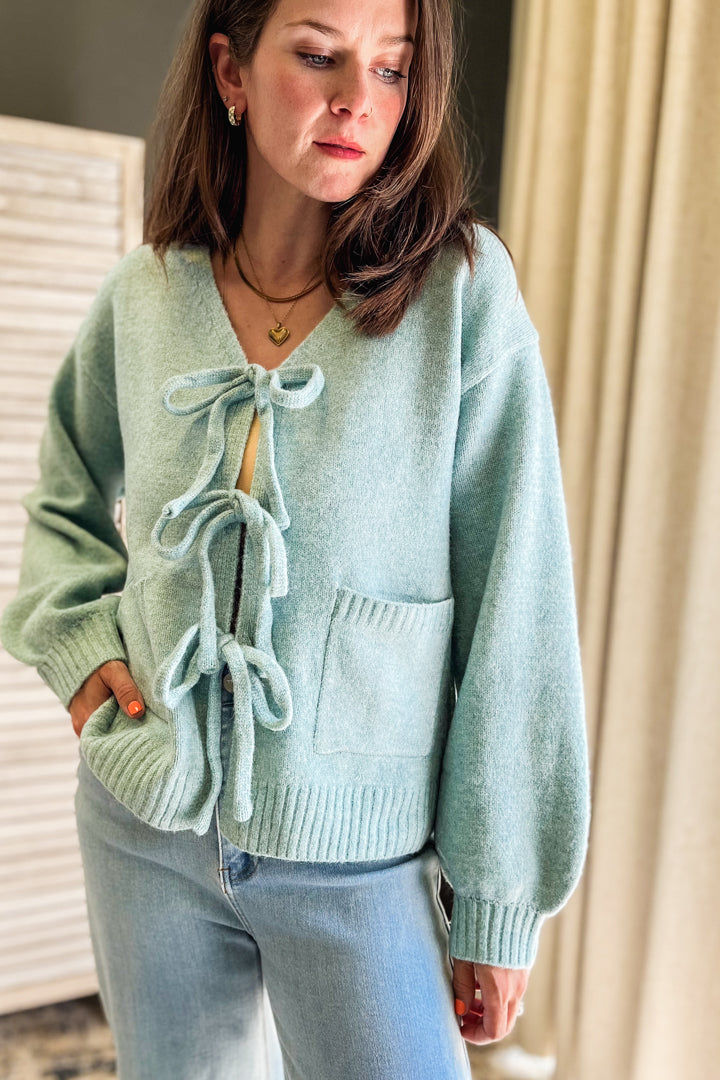 A woman wearing a light mint green knit cardigan sweater with tie-front details and front pockets, paired with light blue jeans in an indoor setting.