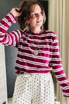 A woman wearing a long-sleeve crewneck shirt with horizontal magenta and white stripes, paired with white pants featuring black polka dots. She is wearing a silver pendant necklace and looking to the side in an indoor setting.