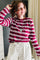 A woman wearing a long-sleeve crewneck shirt with horizontal magenta and white stripes, paired with white pants featuring black polka dots. She is wearing a silver pendant necklace and looking to the side in an indoor setting.