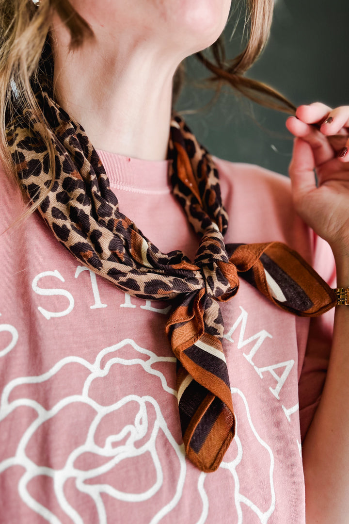 A close-up lifestyle shot of a woman wearing a leopard print and striped silk scarf tied around her neck over a dusty pink graphic t-shirt.