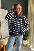 A woman wearing a navy and white striped cardigan with a denim collar and front pockets, paired with light blue jeans, standing in a room with a dark gray wall and a potted plant.