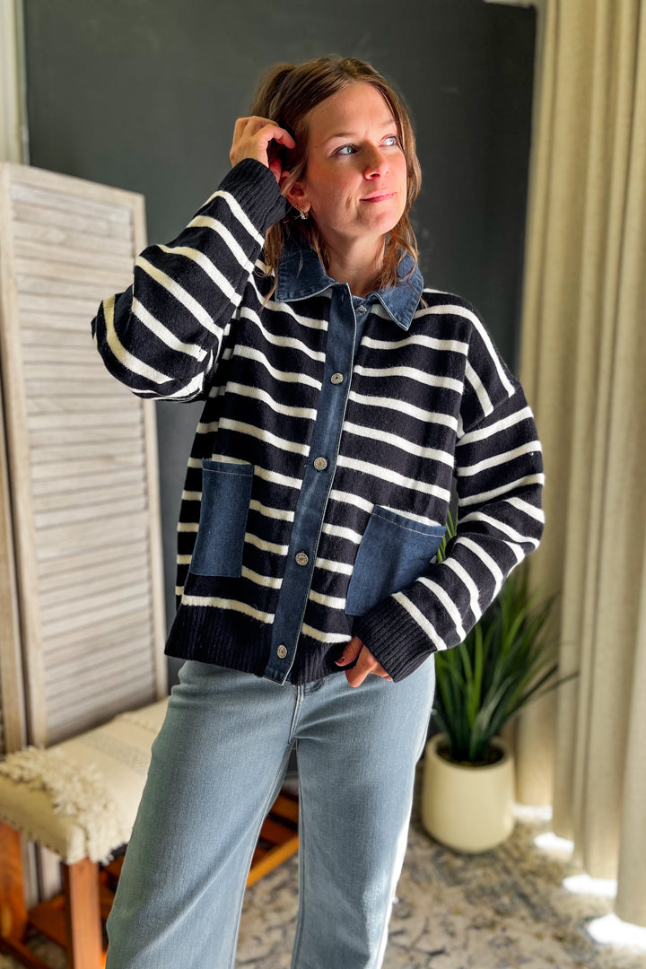 A woman wearing a navy and white striped cardigan with a denim collar and front pockets, paired with light blue jeans, standing in a room with a dark gray wall and a potted plant.