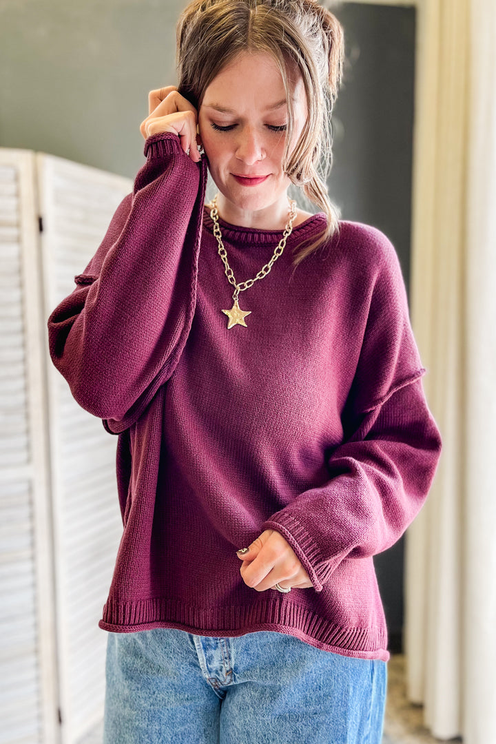 A woman wearing a loose-fitting, long-sleeve crewneck sweater in a deep plum or burgundy shade, paired with blue jeans and a gold star pendant necklace, standing indoors.