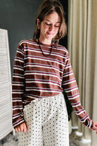 A woman wearing a brown long-sleeve striped shirt with thin red and white stripes, paired with white pants featuring black polka dots, standing in an indoor setting.