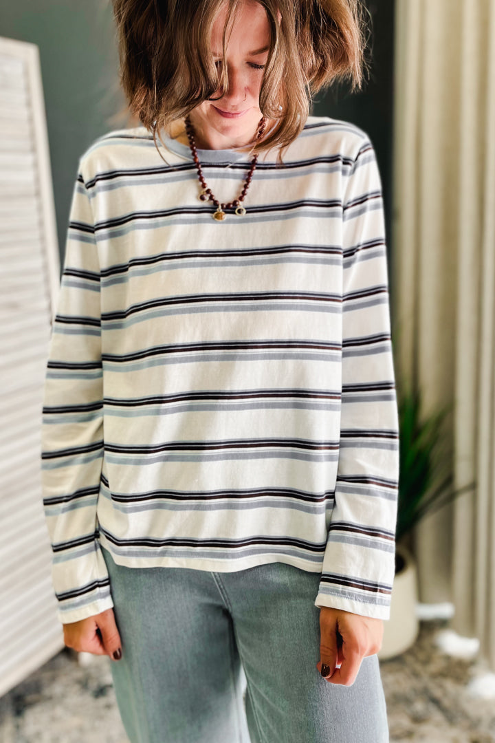 A woman wearing a cream-colored long-sleeve shirt with thin navy and gray horizontal stripes, paired with light blue jeans and a beaded necklace.
