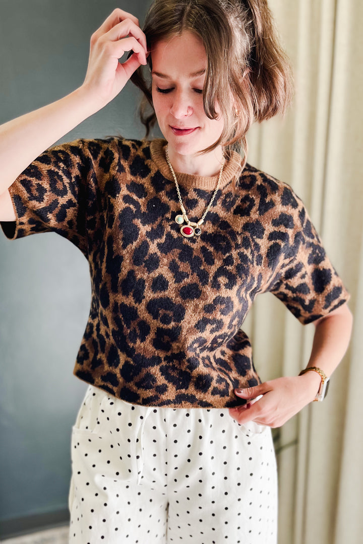 A woman wearing a short-sleeve leopard-print knit top and white pants with black polka dots, standing indoors against a gray wall and beige curtain background.