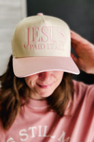 A person wearing a beige baseball cap with pink embroidered text reading 'JESUS PAID IT ALL' and a pink sweatshirt, with the cap covering most of their face.