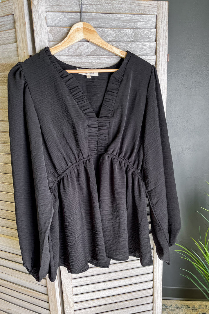A black long-sleeve blouse with a gathered V-neckline and peplum waist, hanging on a wooden hanger against a rustic gray wooden shutter door.