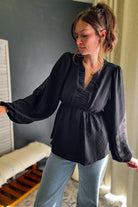 A woman wearing a black long-sleeve blouse with a gathered ruffle neckline and waist, paired with light blue jeans, standing in an indoor setting.