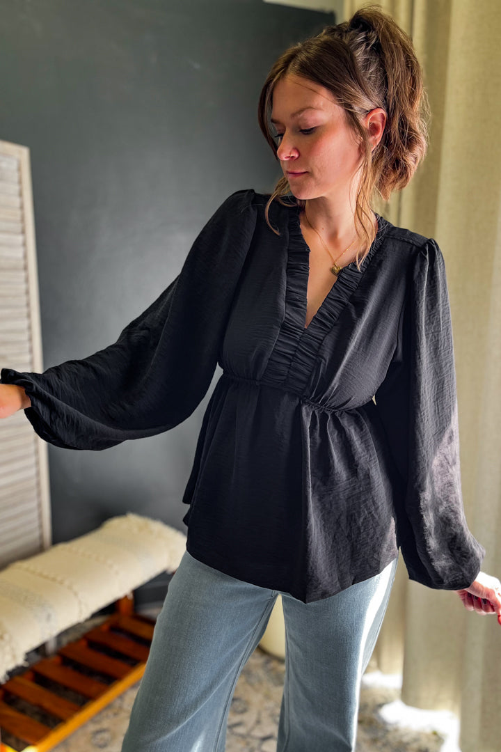 A woman wearing a black long-sleeve blouse with a gathered ruffle neckline and waist, paired with light blue jeans, standing in an indoor setting.
