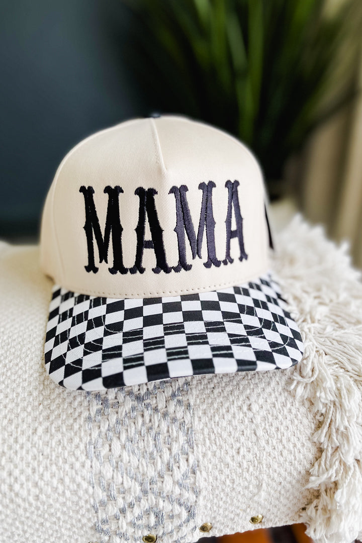 A cream-colored baseball cap with 'MAMA' embroidered in black across the front and a black-and-white checkered pattern on the visor, resting on a textured cream throw blanket.