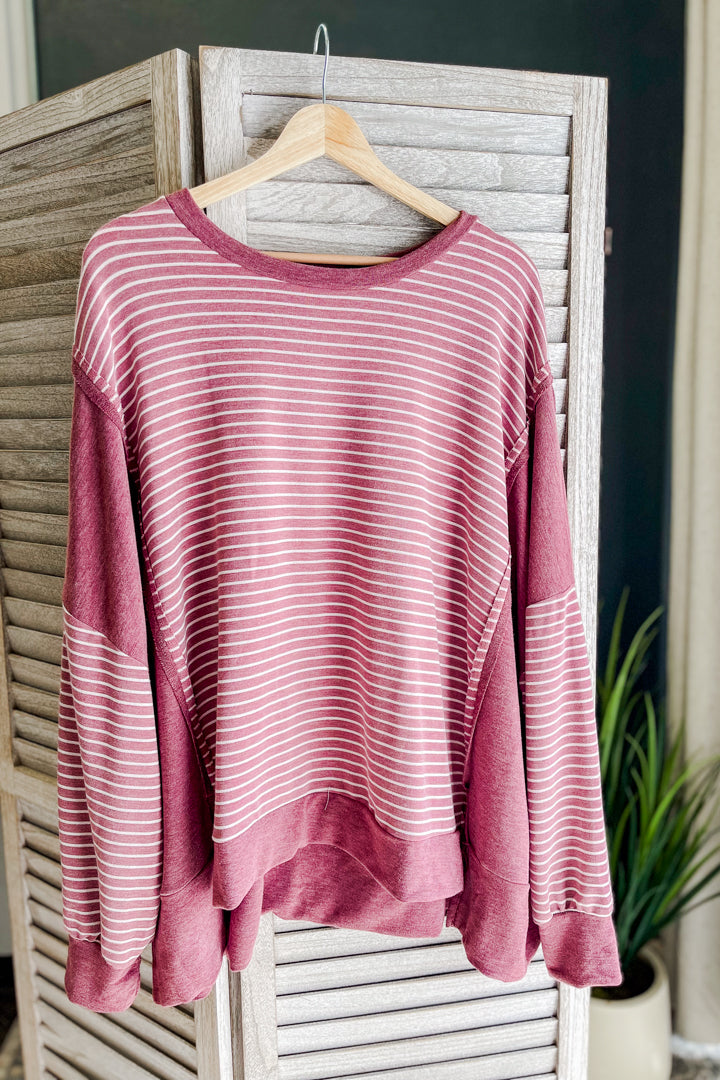 A dusty rose long-sleeve top with thin white horizontal stripes, hanging on a wooden hanger against a rustic white slatted screen with a green plant in the background.