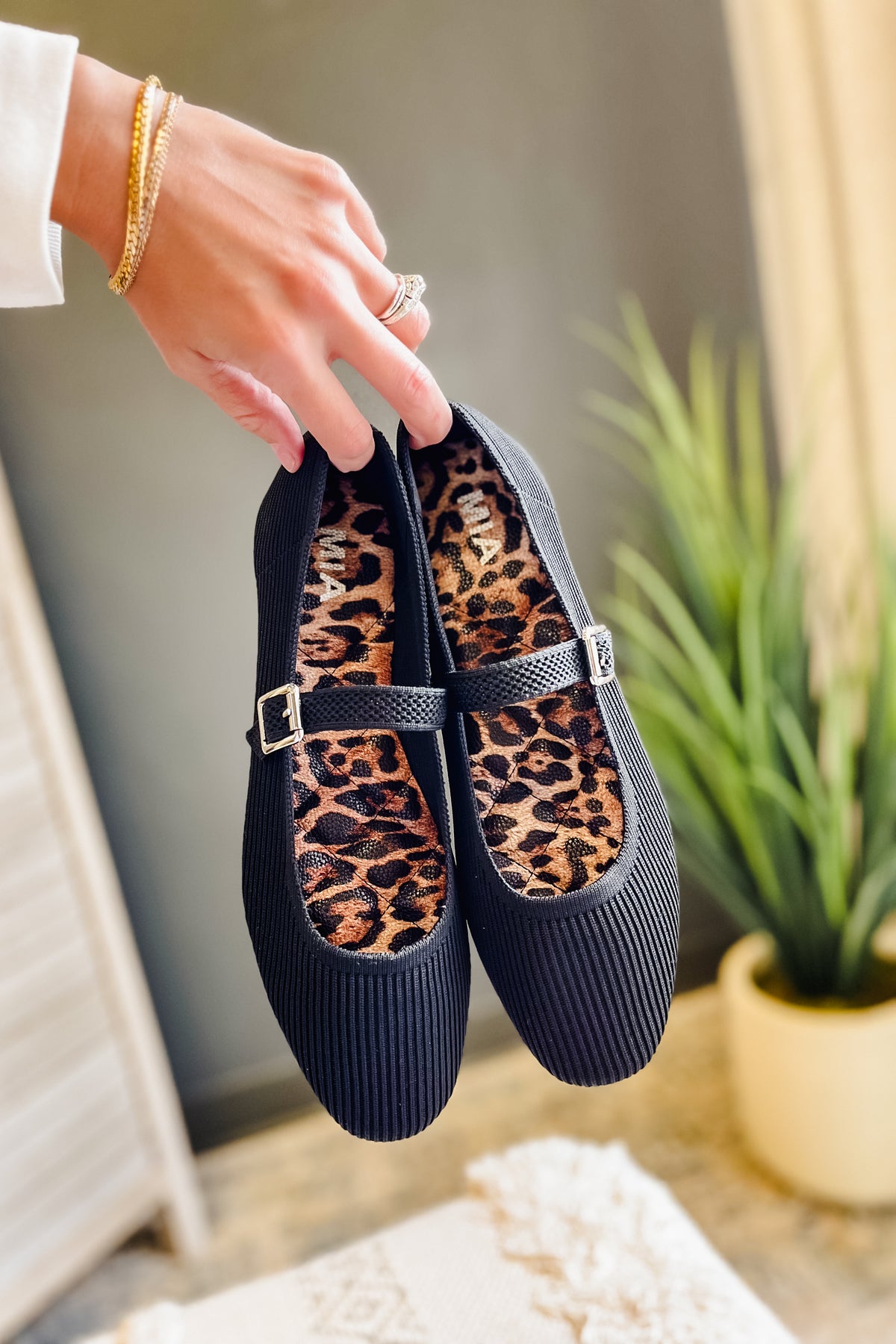 A pair of black ribbed Mary Jane flats with leopard print interiors, held by a hand with a gold bracelet in a home setting with a potted plant in the background.