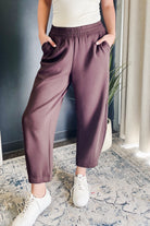 A person wearing loose, wide-leg cropped pants in a dusty mauve or plum-brown color, paired with a white top and white sneakers, standing indoors on a patterned rug.