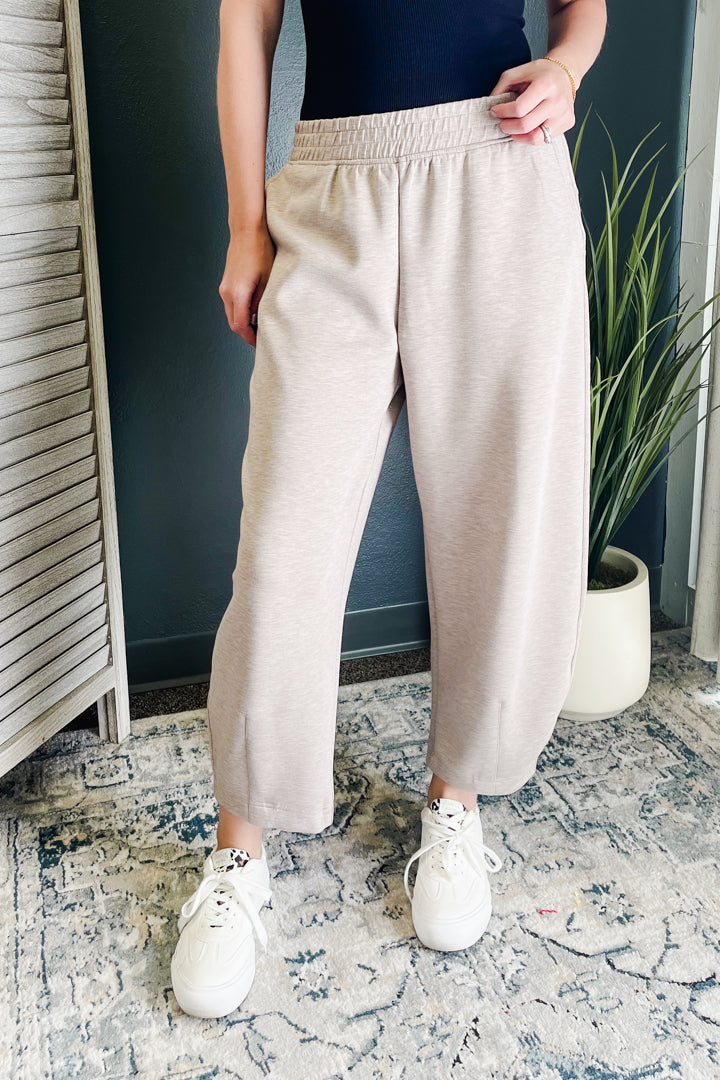 A person wearing light beige wide-leg cropped sweatpants with an elastic waistband, paired with white sneakers, standing indoors on a patterned rug.