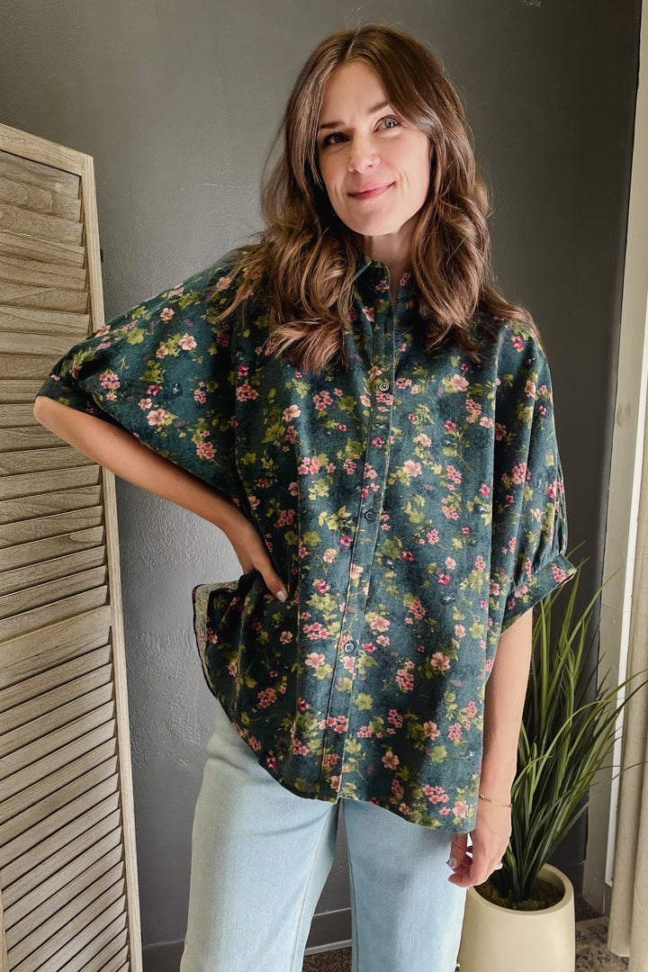 A woman wearing a dark green short-sleeve button-up shirt with a pink and yellow floral pattern, paired with light blue jeans, standing in an indoor setting.