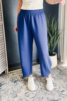 A person wearing loose, wide-leg blue pants with an elastic waistband, paired with white sneakers, standing indoors on a patterned rug near a potted plant.