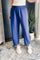 A person wearing loose, wide-leg blue pants with an elastic waistband, paired with white sneakers, standing indoors on a patterned rug near a potted plant.