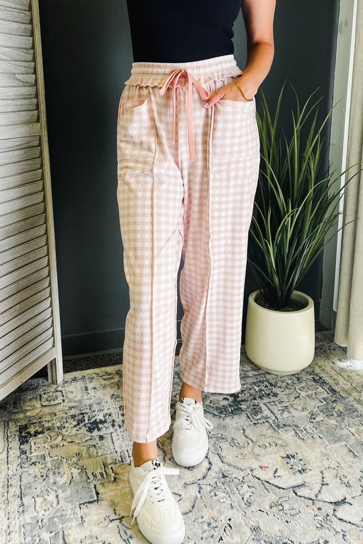 A person wearing pink and white gingham drawstring pants, paired with a black top and white sneakers, standing indoors next to a potted plant.