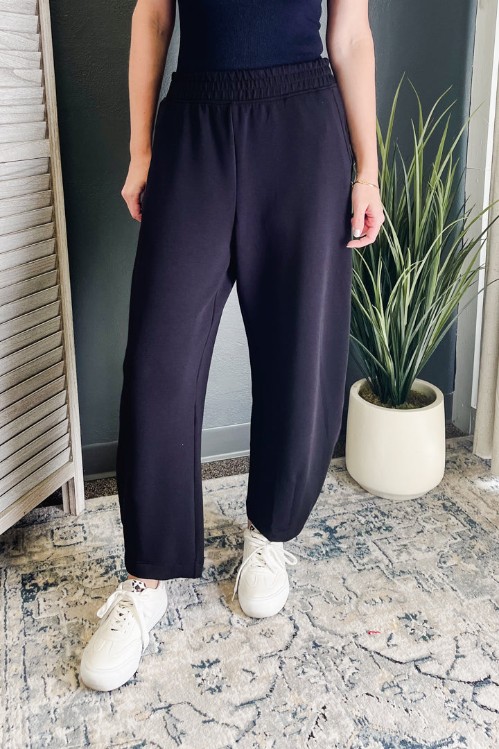 A person wearing dark navy wide-leg sweatpants with an elastic waistband, paired with white sneakers, standing in an indoor setting with a patterned rug and a potted plant.