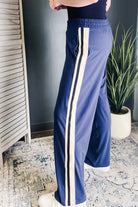 A person wearing blue wide-leg track pants with a double vertical cream stripe down the side, standing indoors near a plant and textured rug.