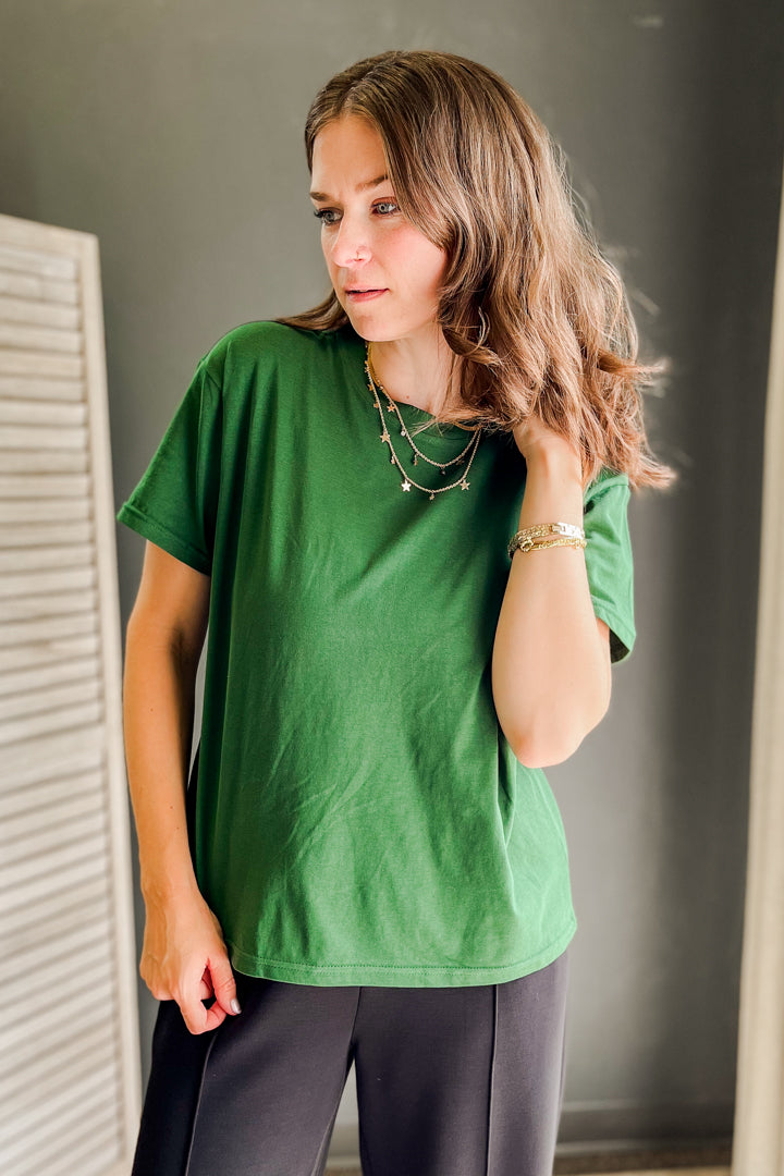 A woman wearing a green short-sleeve t-shirt and dark gray pants, standing indoors against a dark gray wall.
