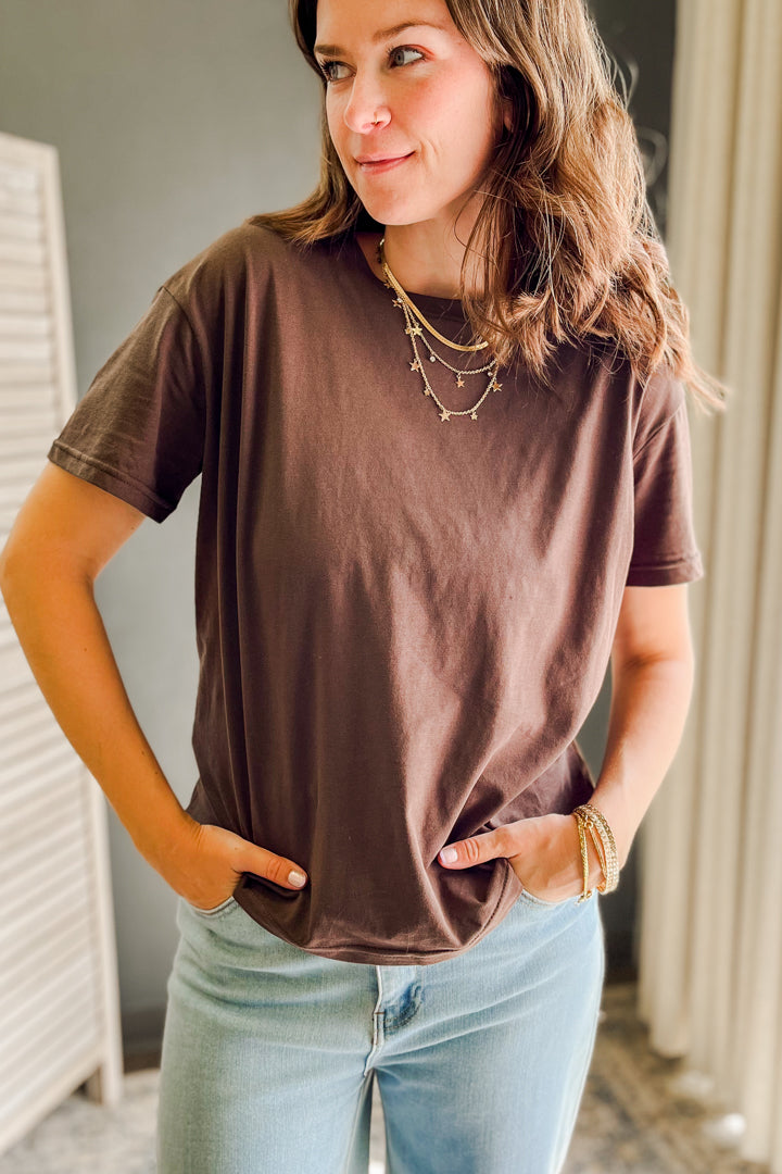 A woman wearing a brown short-sleeve crewneck t-shirt and light blue jeans, standing indoors with her hands in her pockets.