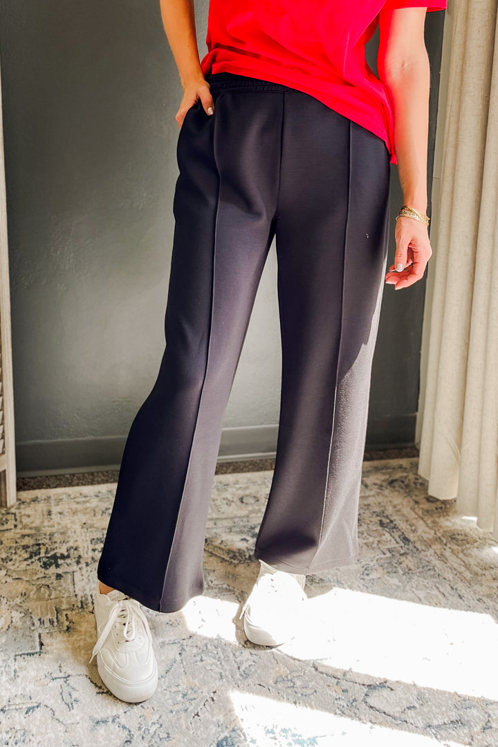 A person wearing dark charcoal wide-leg trousers with a red top and white sneakers, standing indoors on a patterned rug.