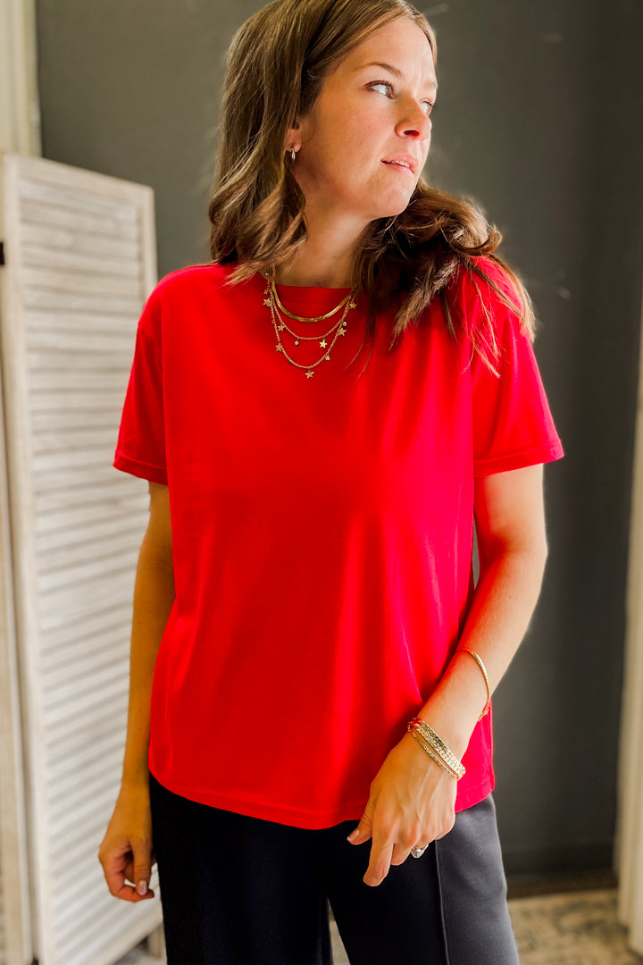 A woman wearing a bright red short-sleeve crewneck t-shirt and dark pants, standing in an indoor setting with a dark gray wall and a white louvered door in the background.