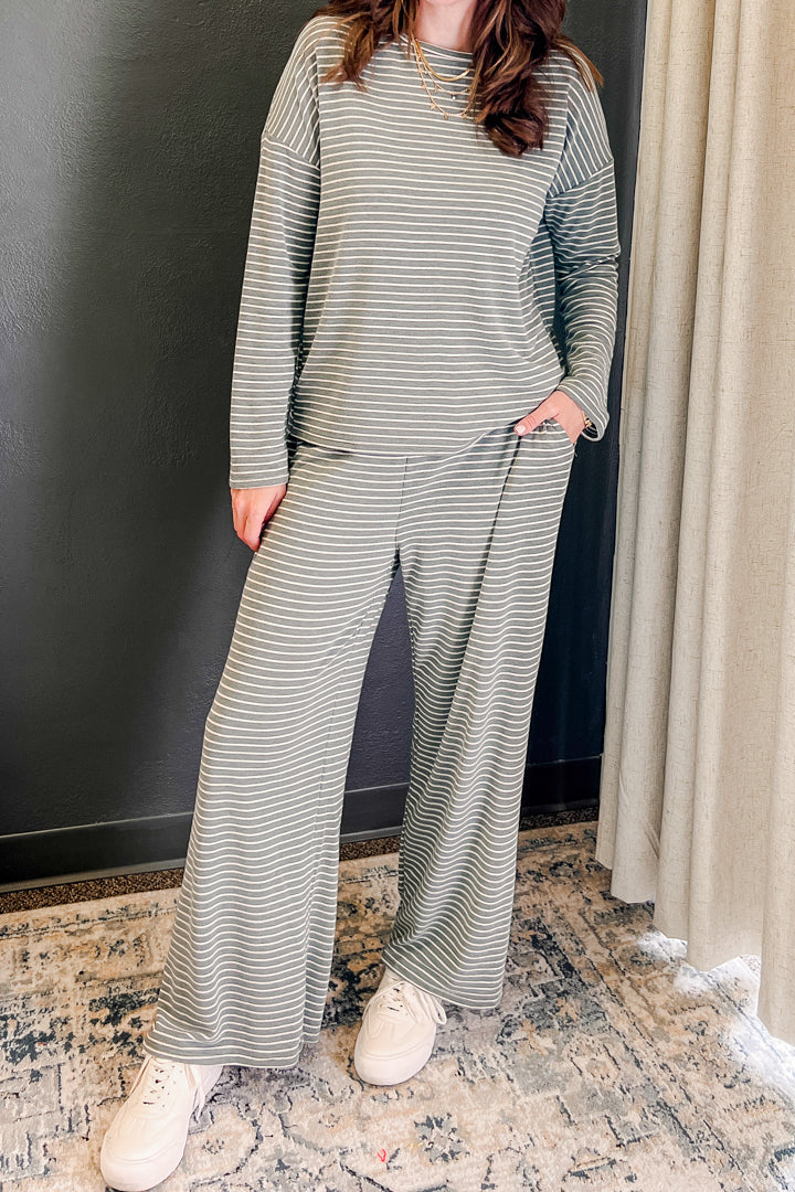 A woman wearing a matching two-piece loungewear set with a muted green and white horizontal stripe pattern, featuring a long-sleeve top and wide-leg pants, paired with white sneakers.