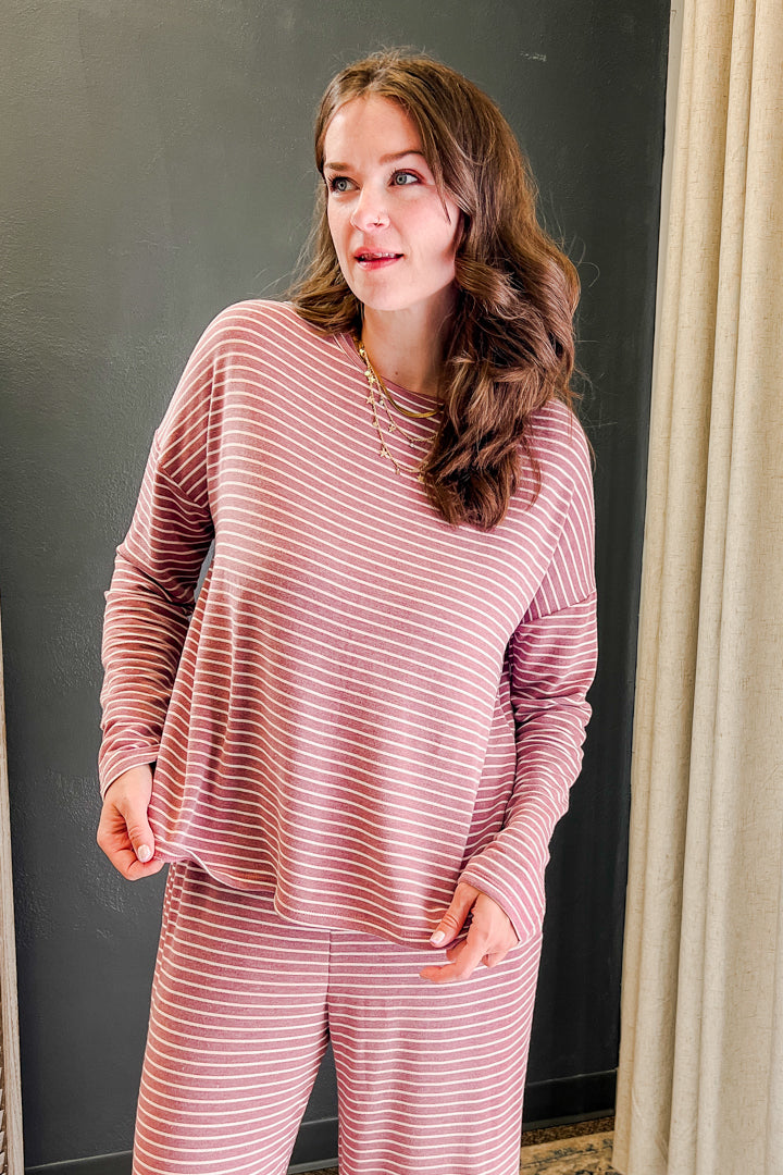 A woman wearing a matching two-piece pajama set with a dusty rose and white striped pattern, standing in front of a dark gray wall and a beige curtain.