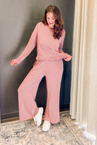 A woman modeling a matching two-piece loungewear set consisting of a long-sleeve wrap-style top and wide-leg pants, both in a dusty pink and white striped pattern. She is standing indoors next to a dark gray wall and a beige curtain.