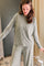 A woman wearing a matching two-piece loungewear set with a long-sleeve top and wide-leg pants, both in a muted sage green with thin white horizontal stripes. She is standing indoors next to a beige curtain.
